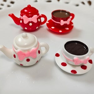 May include: Two miniature tea sets featuring a red teapot with white polka dots and a white teapot with red polka dots. Each set includes a matching teacup on a saucer filled with dark brown liquid, and each piece is decorated with a small pink bow.