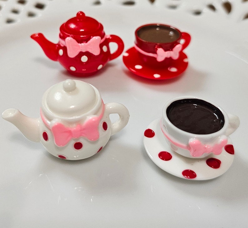 May include: Two miniature tea sets featuring a red teapot with white polka dots and a white teapot with red polka dots. Each set includes a matching teacup on a saucer filled with dark brown liquid, and each piece is decorated with a small pink bow.