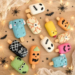 May include: A collection of Halloween-themed popsicles in various shapes and colors, including ghosts, pumpkins, and monsters. The popsicles are arranged on a wooden surface with decorative spiders and bats.