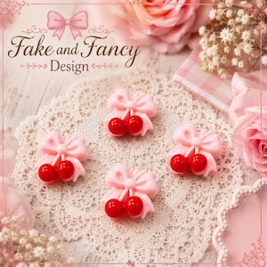 May include: Four small, three-dimensional cherry-shaped embellishments, each featuring a pair of glossy red cherries attached to a light pink bow. The items are arranged on a white lace doily, with pink roses and baby's breath flowers visible.