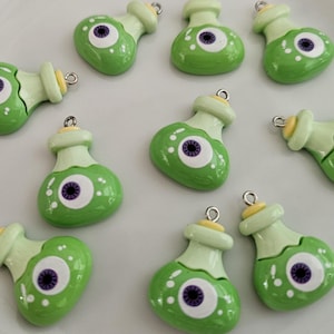 May include: Green potion bottle charms with a large eye design. The charms have a light green body, a light green neck, and a yellow cap. Each charm has a silver loop for attaching to jewelry or other crafts.