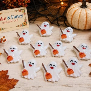 May include: A collection of ten small white ghost figurines, each featuring a smiling face and holding a miniature orange broom. The ghosts are arranged in a group, and a rectangular card with the text Fake & Fancy DESIGN LLC is visible.