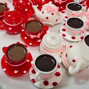 May include: A collection of miniature resin tea sets featuring red and white polka dot patterns. The set includes small teapots and teacups filled with dark brown liquid, accented with small pink bows on the handles and lids of the pieces.