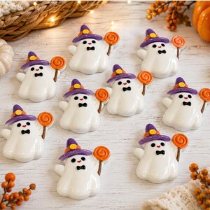 May include: A collection of nine small, white ghost figurines, each wearing a purple witch hat with a yellow band and holding an orange lollipop. The ghosts have black eyes, a small red mouth, and a black bowtie, arranged on a light surface.