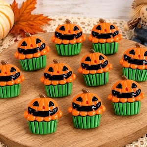 May include: A collection of ten small cupcake-shaped ornaments, each featuring an orange jack-o'-lantern topper with a black carved face and a green fluted base. These miniature decorative items are arranged on a round wooden surface.