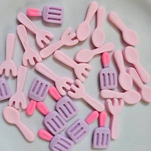 May include: A collection of pastel-colored miniature food-themed embellishments. The assortment includes pink forks, spoons, and popsicle-shaped charms with pink and purple accents. These small decorative pieces are scattered on a white surface.