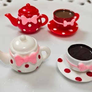 May include: Two miniature tea sets featuring a red teapot with white polka dots and a white teapot with red polka dots. Each set includes a matching cup and saucer, both adorned with a small pink bow detail and filled with a dark brown liquid.