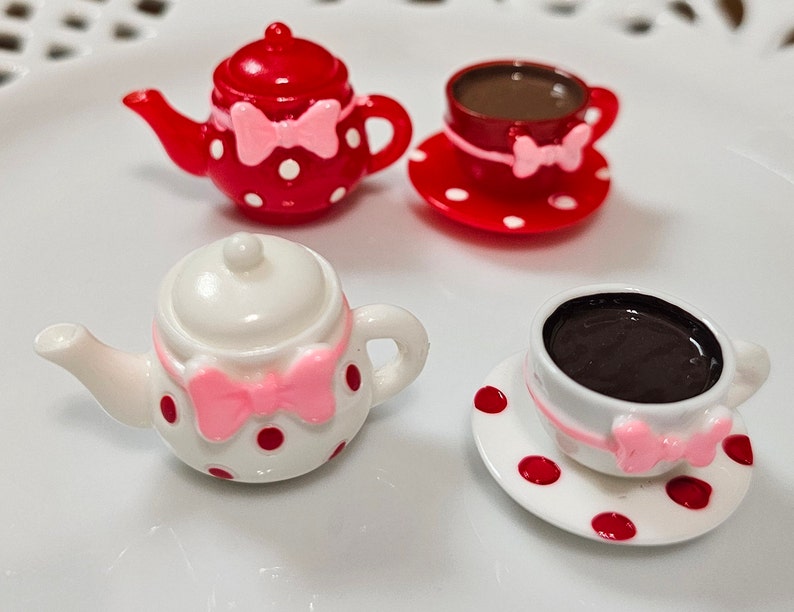 May include: Two miniature tea sets featuring a red teapot with white polka dots and a white teapot with red polka dots. Each set includes a matching cup and saucer, both adorned with a small pink bow detail and filled with a dark brown liquid.