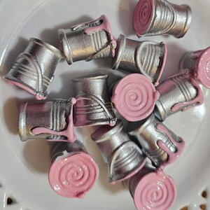 May include: A collection of miniature paint cans with silver and pink accents. The cans are arranged in a circular pattern on a white surface.