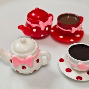 May include: A miniature tea set consisting of two teapots and two teacups with saucers. One teapot and cup set is red with white polka dots, while the other is white with red polka dots. Each piece features a small pink bow detail.