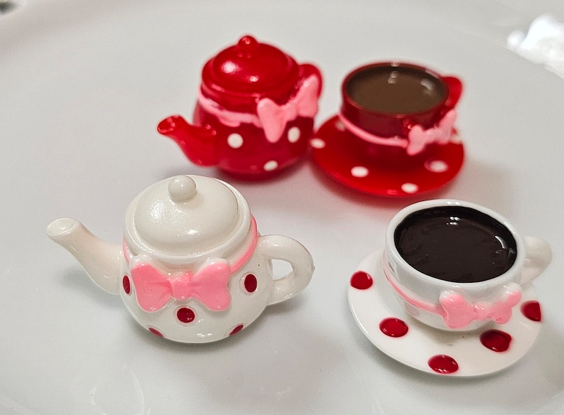 May include: A miniature tea set consisting of two teapots and two teacups with saucers. One teapot and cup set is red with white polka dots, while the other is white with red polka dots. Each piece features a small pink bow detail.