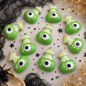 May include: Ten green potion bottle charms, each featuring a central white eyeball with a purple iris and pupil. The charms have a light green neck, a yellow stopper top, and a small silver metal loop for hanging, arranged in a scattered pattern.