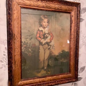 Puede incluir: Retrato enmarcado de un niño pequeño sosteniendo un perro pequeño. El retrato está en un marco de madera marrón, decorativo y ornamentado. El niño lleva una chaqueta roja y pantalones color canela. El fondo presenta una escena de jardín.