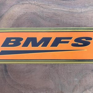 May include: A rectangular sticker featuring a vibrant orange background with the black text BMFS centered in the middle. The design includes a horizontal black underline beneath the letters and three thin black stripes along the top and bottom edges.