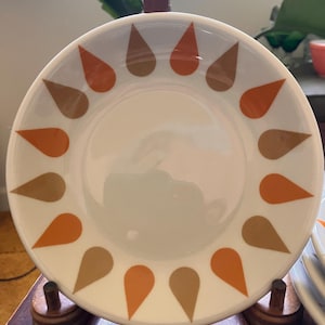 May include: A white ceramic saucer with a brown and orange teardrop pattern. The saucer is on a wooden stand.