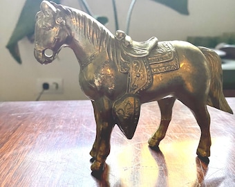 May include: A detailed brass horse figurine with a saddle and shield. The horse is posed in a standing position, showcasing intricate details. The figurine is a warm gold color, suggesting an antique or vintage aesthetic. The horse is on a wooden surface.