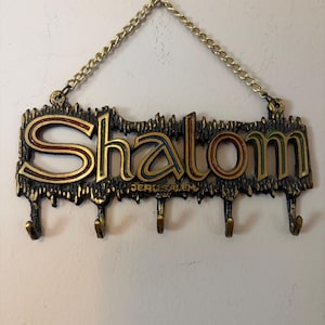 May include: A decorative metal key holder with the word "Shalom" in gold, red, and blue lettering. The word "Jerusalem" is below the word "Shalom". The key holder has four hooks and a gold chain for hanging.