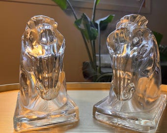 May include: Two clear glass bookends shaped like horse heads. The bookends are on a gold surface.
