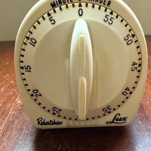 May include: An off-white kitchen timer with a central rotating knob. The face features black numerals from 0 to 55, with the text "MINUTE MINDER" at the top, and "Robertshaw" and "Lux" printed in black script at the bottom of the rounded casing.
