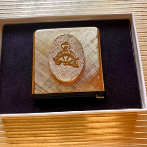May include: A gold-toned metal lighter with a textured surface, presented in a white box with a black interior. The lighter features an oval emblem with a nautical design of a person at a ship's wheel.