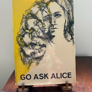 May include: A hardcover book titled "GO ASK ALICE" with a yellow cover and a black and white illustration of a woman's face. The book is propped up on a wooden stand, showcasing the title.