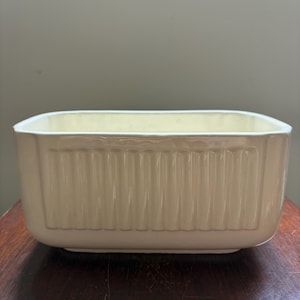May include: Cream-colored rectangular ceramic baking dish. The dish has a ribbed design on the sides and a smooth interior. The dish is sitting on a dark wooden surface.