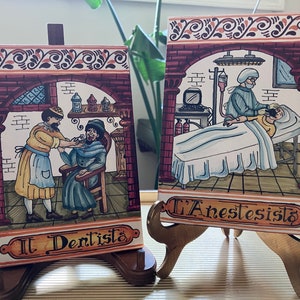 May include: Two colorful ceramic tiles depicting scenes from a dentist's office and an operating room. The dentist tile shows a dentist working on a patient's teeth. The operating room tile shows a surgeon operating on a patient. Both tiles have the words "Il Dentista" and "L'Anestesista" written on them.