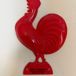 May include: A bright red plastic rooster figurine. The rooster has a detailed comb, wattle, and tail feathers. The base of the figurine has the text "BELL'S MARKET PHONE EM 2-8311 ERWIN S. DAK."