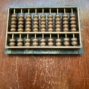 May include: A rectangular abacus featuring a dark green marble base and a metallic frame. The device contains nine vertical rods with multiple polished bronze-colored beads arranged in two sections, separated by a horizontal metal bar.