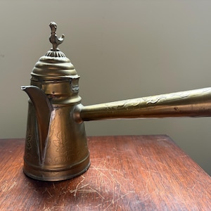May include: An antique brass coffee pot with a long, ornate handle and a decorative bird finial. The pot features etched floral patterns and a spout for pouring. It sits on a wooden surface.
