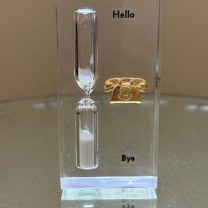 May include: A clear rectangular acrylic block containing a glass hourglass with white sand. To the right of the hourglass is a small gold-colored rotary telephone charm. The word Hello is printed above the charm, and the word Bye is printed below.