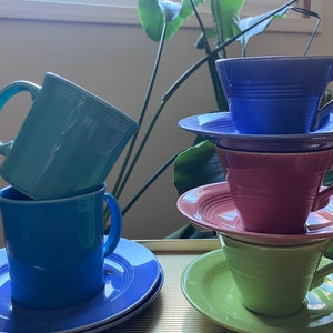 May include: A stack of four ceramic teacups and saucers in various colors, including blue, green, pink, and light blue. The cups and saucers are stacked on a table with a light wood grain pattern.