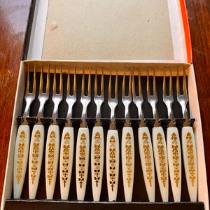 May include: A set of vintage dessert forks in a box. The forks have white handles with gold floral designs. The tines are silver. The box is black with a white interior and a red and orange trim.
