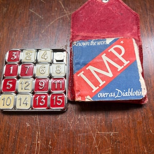 May include: A vintage 15-puzzle game with red and cream numbered tiles in a metal frame, accompanied by a red leather pouch containing a square instruction booklet with the text IMP, Known the world, and over as Diablotin printed on the cover.