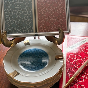 May include: A vintage set featuring playing cards, an ashtray, and a score pad. The playing cards are in a clear plastic case and have a floral pattern. The ashtray is white with gold trim and a blue floral center. The score pad is red.