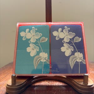 May include: A vintage Hallmark playing card set in a clear plastic case with a red frame. The cards feature a floral design with white flowers on a teal and navy blue background. The set is displayed on a wooden stand.