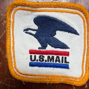 May include: A square embroidered patch with a white background and a thick yellow border. The center features a dark blue eagle silhouette above a red horizontal stripe, the text U.S.MAIL in black, and a blue horizontal stripe at the bottom.
