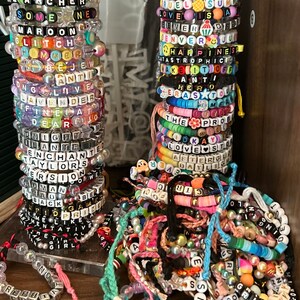 May include: A collection of colourful beaded bracelets with various words and phrases, including "HE ARCHER", "SOME MAROON", "GLITCH", "LOVE IS A", "HAPPINESS", "TAKETOFF", "HERO", "THE PROB", "LOVE STO", "AFTERGLE", "DAYLIGH", and more. The bracelets are stacked on two clear acrylic stands.