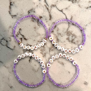 May include: Four beaded bracelets with purple beads and white letter beads spelling out "LOONATIC", "HOLLANOV", "HOLLANDER", and "ROZANOV". The bracelets have gold-colored accents.