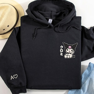 Puede incluir: Sudadera con capucha negra con un coraz&oacute;n blanco y el texto "A" en la manga izquierda. La parte delantera de la sudadera presenta un personaje de dibujos animados en blanco y negro con el texto "クロミ" en japon&eacute;s.