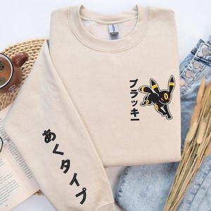 May include: Beige sweatshirt with a black and yellow cartoon character design and Japanese text. The sleeve features additional Japanese text. The sweatshirt is paired with blue jeans, a cup of coffee, and a book with glasses.