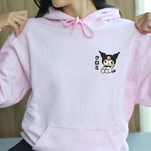 Puede incluir: Una sudadera con capucha rosa con un gr&aacute;fico en blanco y negro de un personaje de dibujos animados con el texto "クロミ" en la parte delantera. La sudadera tiene un coraz&oacute;n bordado en la manga izquierda.