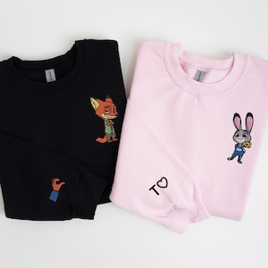 May include: Two black and pink sweatshirts with cartoon animal characters. The black sweatshirt has a fox character on the front and a hand giving a thumbs up on the sleeve. The pink sweatshirt has a bunny character on the front and the text "T♡" on the sleeve.