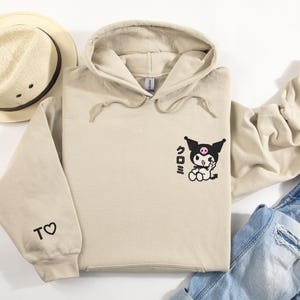 Puede incluir: Una sudadera con capucha beige con un personaje de dibujos animados negro y rosa en la parte delantera. El personaje est&aacute; haciendo un signo de paz y tiene la palabra japonesa "Kuromi" escrita a su lado. La sudadera tiene un peque&ntilde;o coraz&oacute;n con la letra "T" en la manga izquierda.