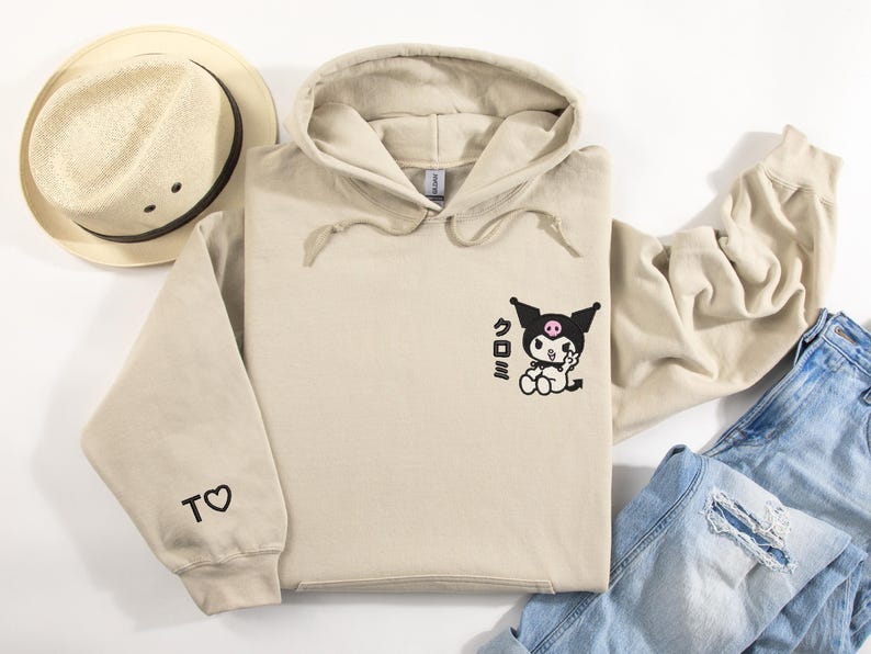 Puede incluir: Una sudadera con capucha beige con un personaje de dibujos animados negro y rosa en la parte delantera. El personaje est&aacute; haciendo un signo de paz y tiene la palabra japonesa "Kuromi" escrita a su lado. La sudadera tiene un peque&ntilde;o coraz&oacute;n con la letra "T" en la manga izquierda.