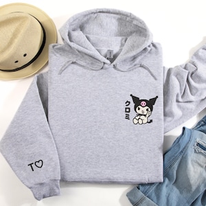 Puede incluir: Una sudadera con capucha gris con un personaje de dibujos animados en blanco y negro en la parte delantera. El personaje lleva un lazo rosa y tiene una calavera negra en la cabeza. El texto "くろみ" est&aacute; impreso debajo del personaje. La sudadera tiene un peque&ntilde;o coraz&oacute;n con una "T" en la manga izquierda.