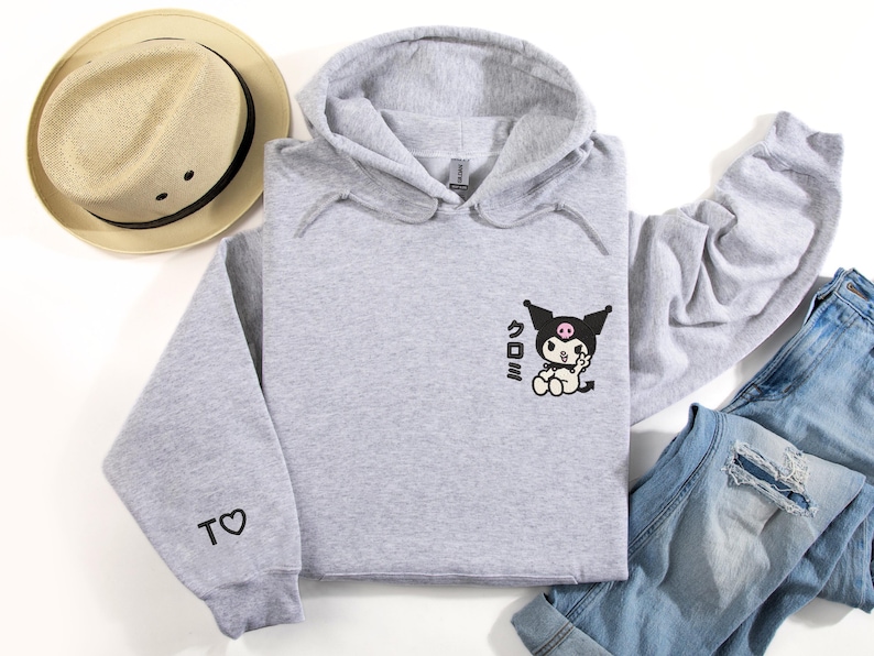 Puede incluir: Una sudadera con capucha gris con un personaje de dibujos animados en blanco y negro en la parte delantera. El personaje lleva un lazo rosa y tiene una calavera negra en la cabeza. El texto "くろみ" est&aacute; impreso debajo del personaje. La sudadera tiene un peque&ntilde;o coraz&oacute;n con una "T" en la manga izquierda.
