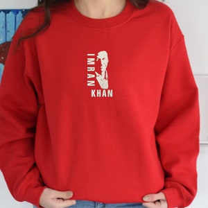 May include: Red sweatshirt with a white graphic of a man's face and the text "IMRAN KHAN" printed on the front.