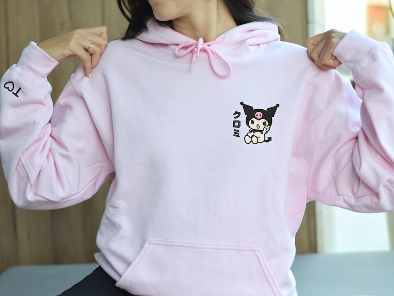Puede incluir: Una sudadera con capucha rosa con un gr&aacute;fico en blanco y negro de un personaje de dibujos animados con el texto "クロミ" en la parte delantera. La sudadera tiene un coraz&oacute;n bordado en la manga izquierda.