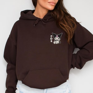Puede incluir: Una sudadera con capucha marr&oacute;n recortada con un gr&aacute;fico en blanco y negro de un personaje de dibujos animados con el texto japon&eacute;s "クロミ" en la parte delantera.